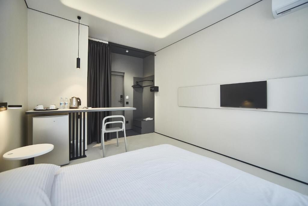 Ein modernes Hotelzimmer mit Bett, Schreibtisch, Stuhl, einer kleinen Küchenzeile und einem an der Wand montierten Fernseher.