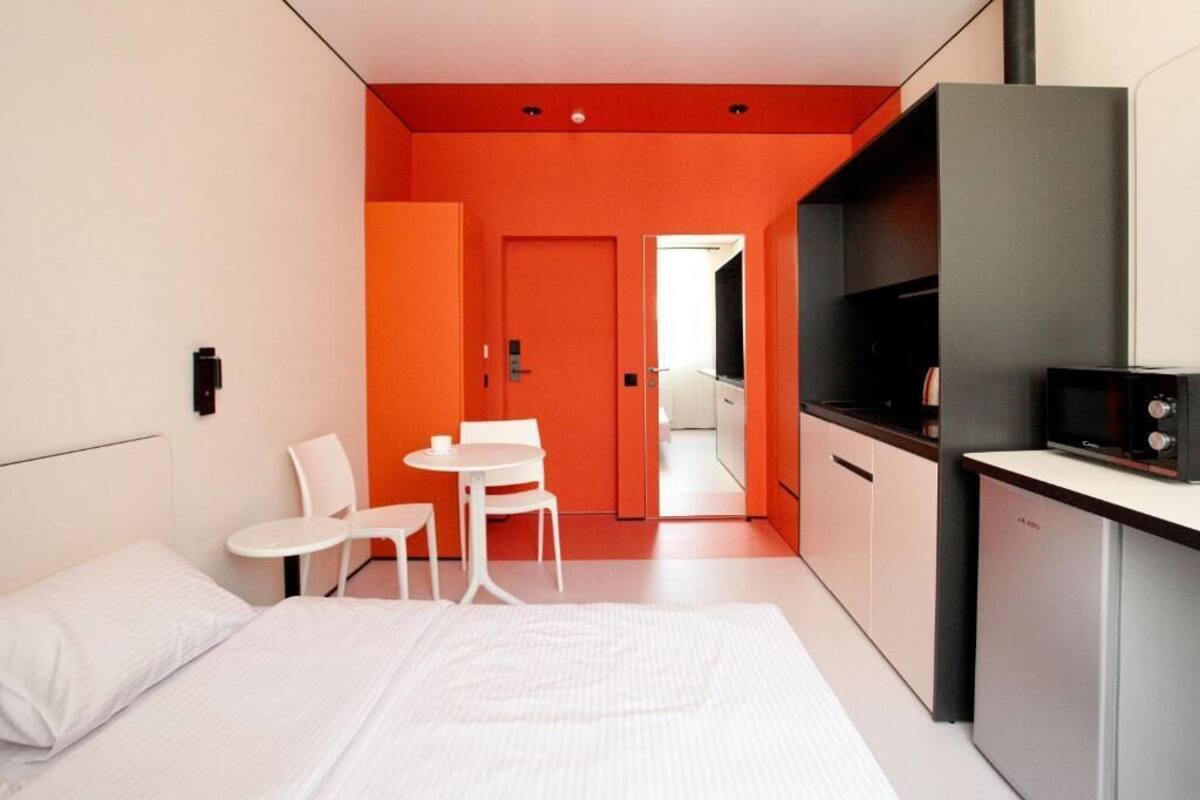Una camera d'albergo moderna con un letto, un piccolo tavolo con sedie, angolo cottura e una porta che dà su un'altra stanza.