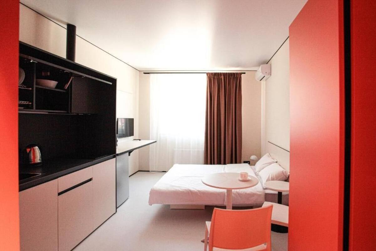 Una camera d'albergo compatta con un letto, un tavolino, una sedia, angolo cottura e una finestra con tende.