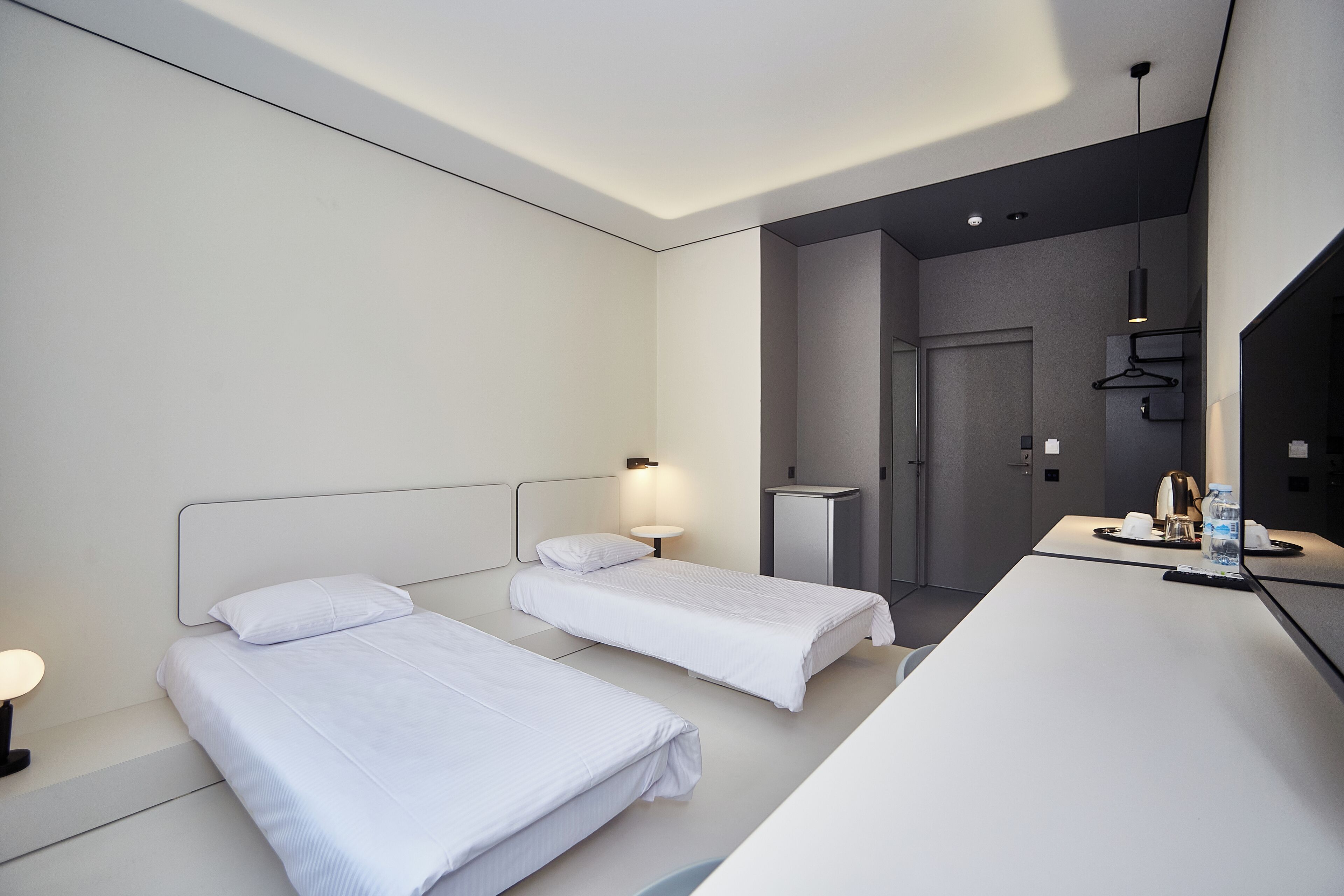 Una habitación de hotel moderna con dos camas individuales, un escritorio, una silla, un espejo y una pequeña kitchenette.