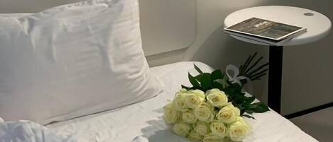 A neatly made bed with white bedding, a white pillow, and a small table with a book and a bouquet of white roses.