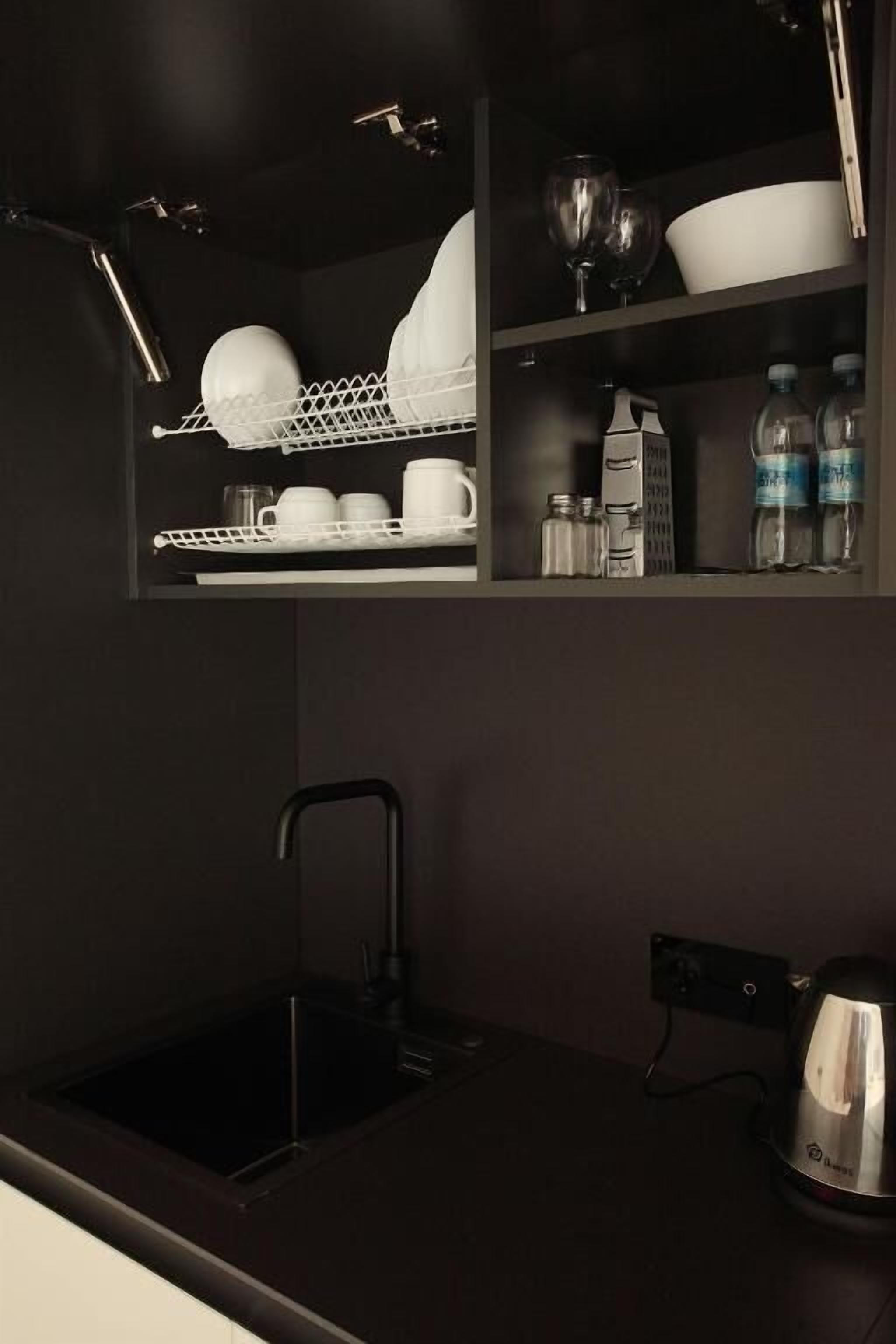 Una cucina con un piano di lavoro nero, un lavello e un bollitore. Sopra il lavello, scaffali con piatti, bicchieri e bottiglie.