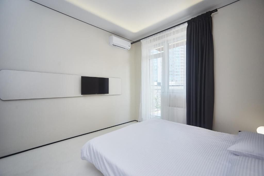 Una habitación de hotel moderna con una cama grande, un televisor montado y un ventanal con cortinas translúcidas.