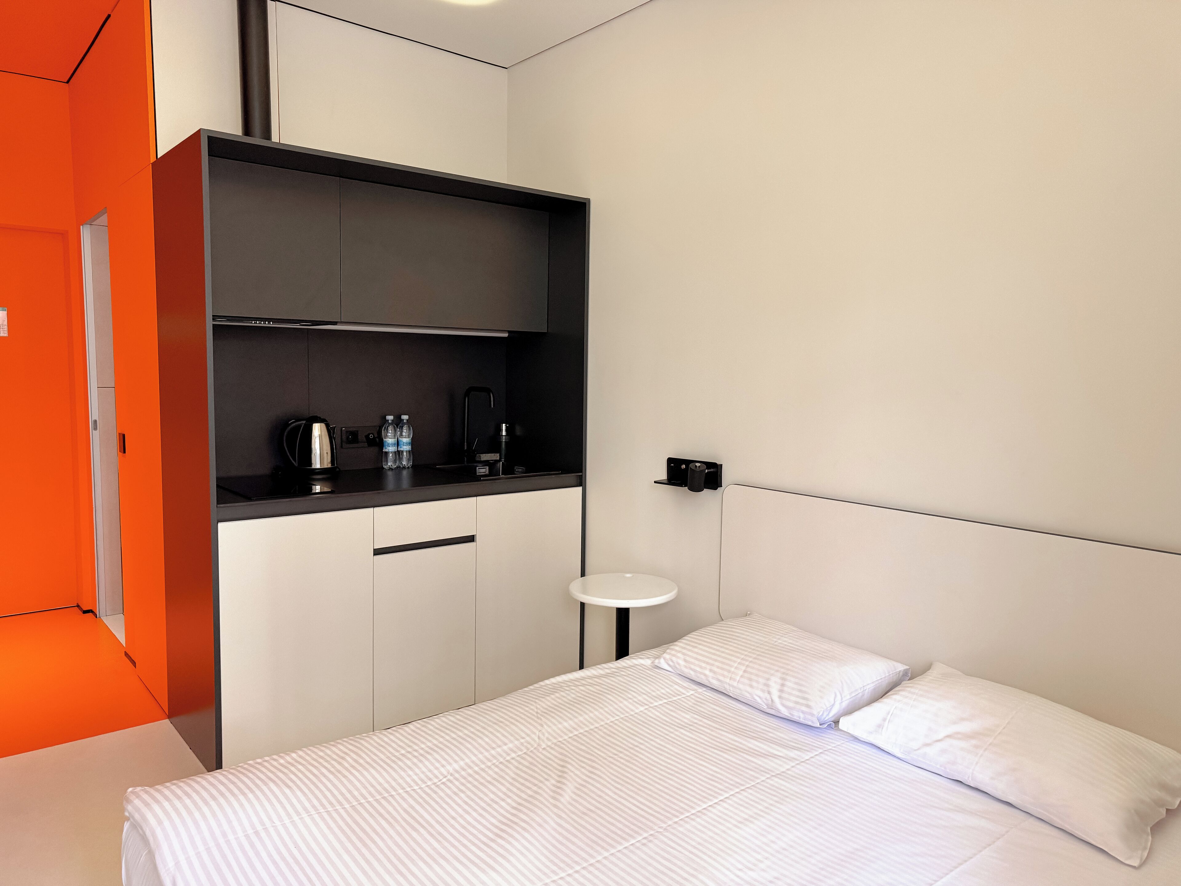 A modern hotel room with a bed, a small bedside table, a built-in kitchen area with a kettle and water bottles, and a bright orange accent wall.