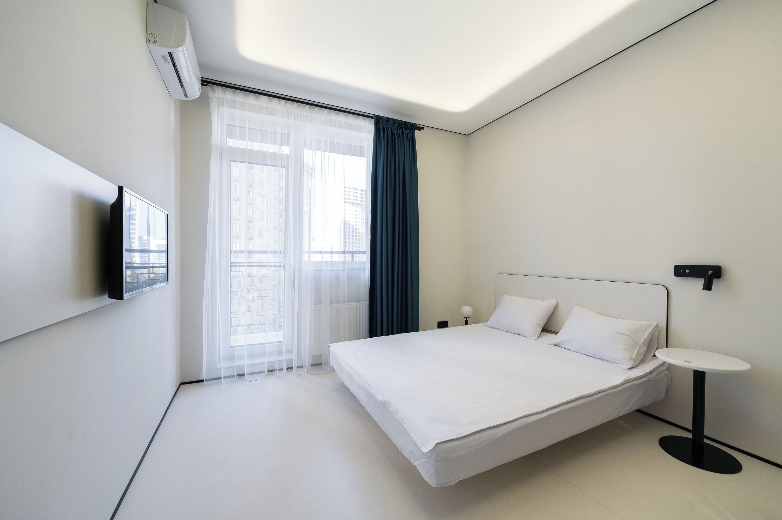 Ein modernes Hotelzimmer mit einem großen Bett, einem kleinen Nachttisch, einem an der Wand befestigten Fernseher und einem Fenster mit durchsichtigen Vorhängen.