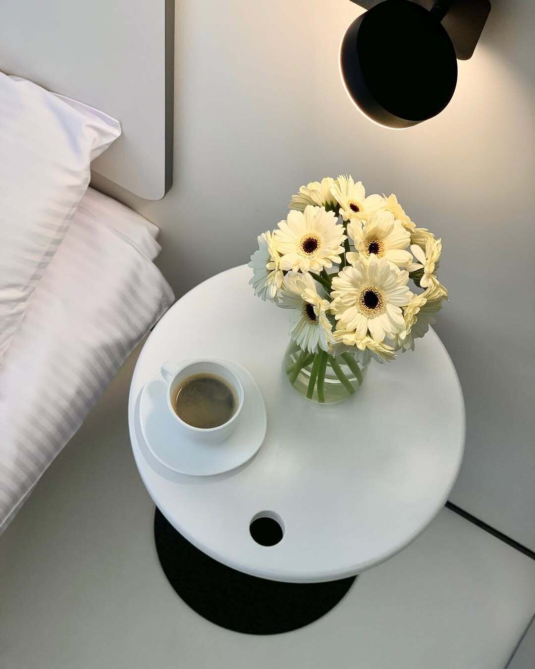 Una mesita de noche blanca con una taza de café blanca, un florero con flores blancas y una lámpara de pared.