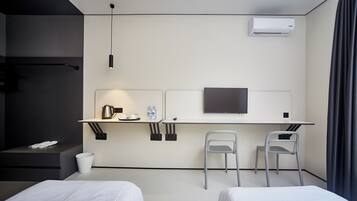 A modern hotel room with a bed, a desk with a flat-screen TV, two chairs, a small table with a kettle and water bottles, and a trash bin.