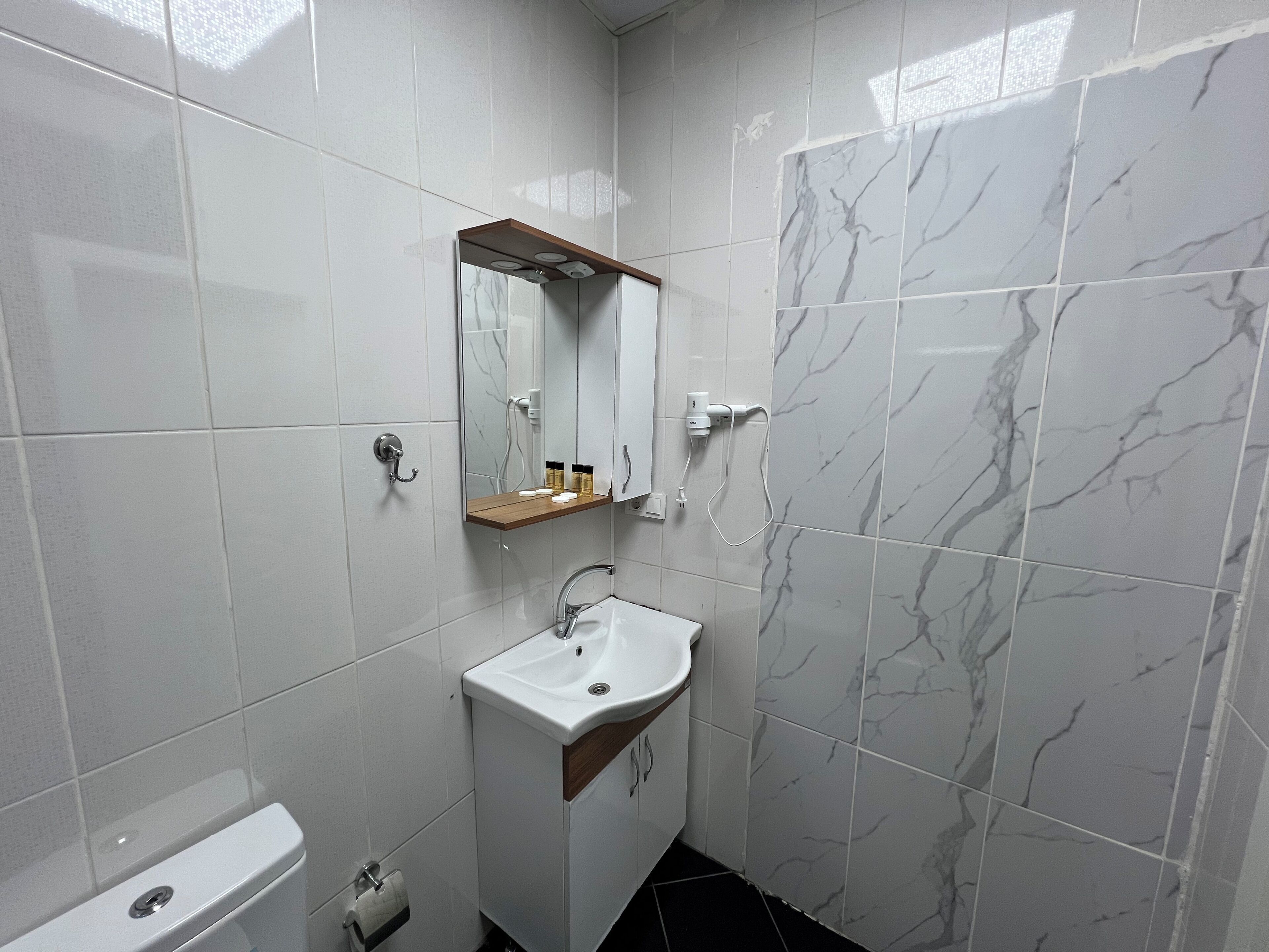Une salle de bain moderne avec des carreaux blancs, un mur imitant le marbre, une étagère en bois au-dessus du lavabo et des toilettes dotées d’un porte-rouleau.