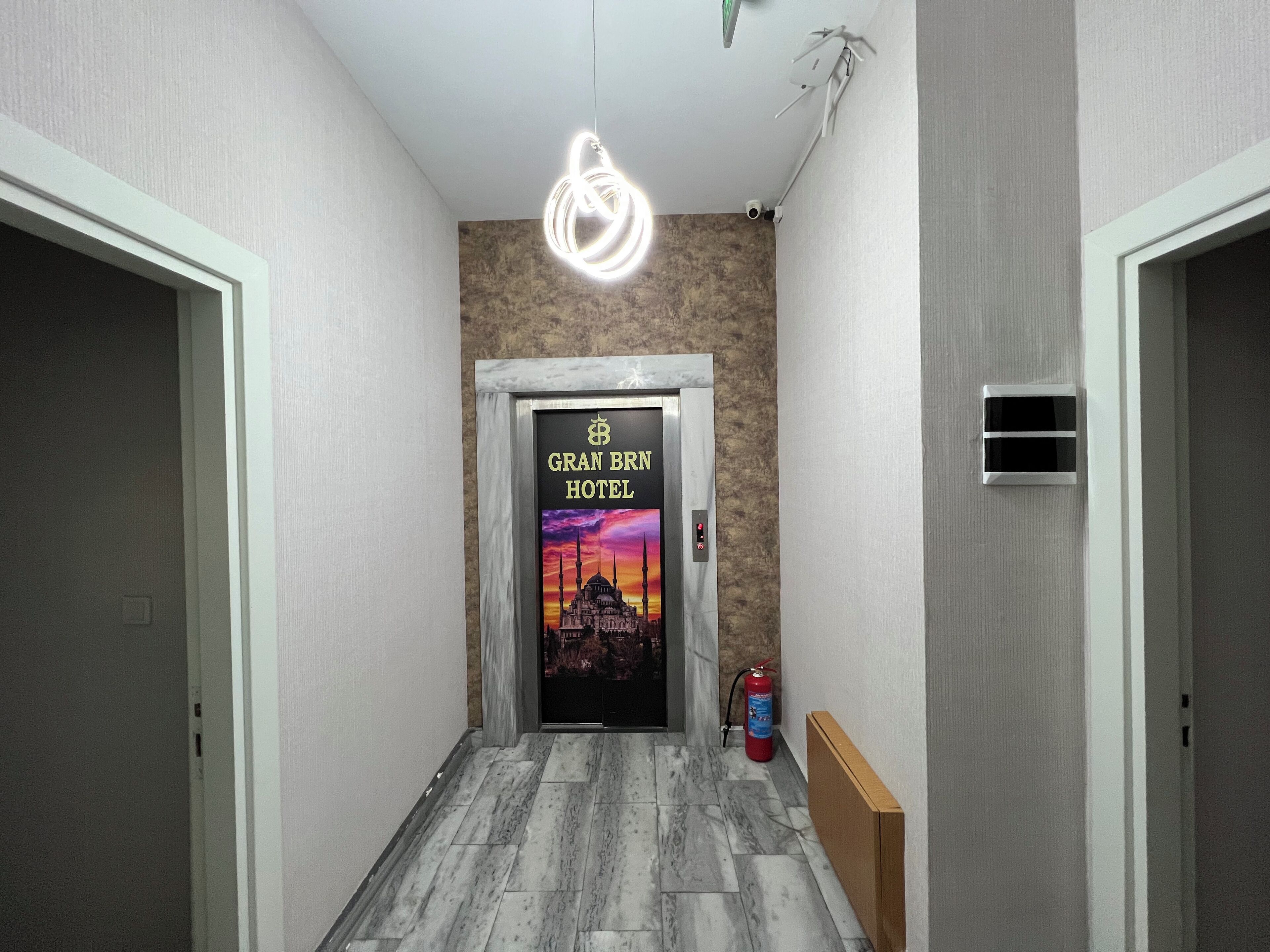 A hallway with a framed sign for GRAN BRN HOTEL, a fire extinguisher, and a ceiling light fixture.