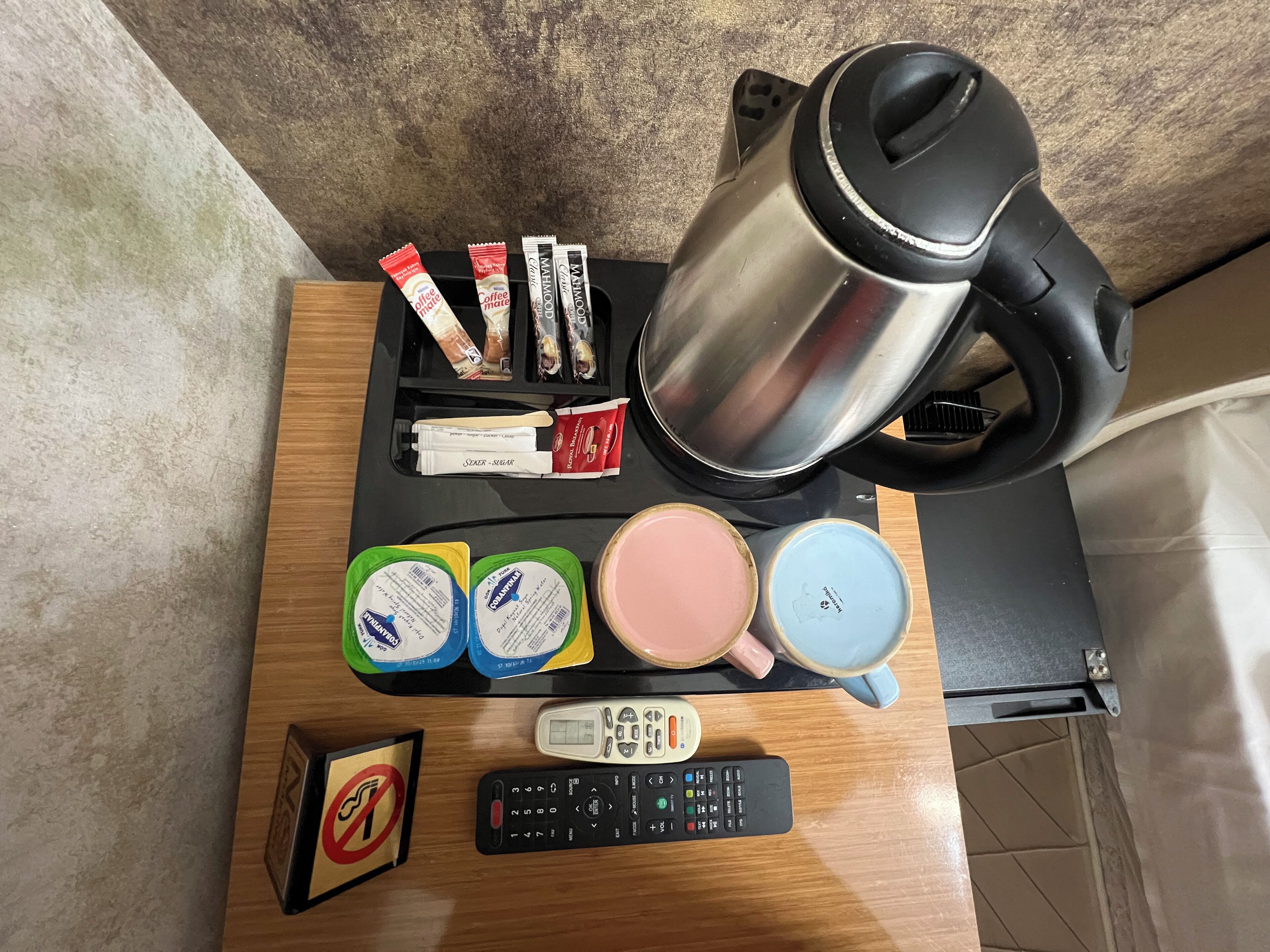 A wooden table with a coffee maker, tea bags, a thermos, a pink mug, a blue mug, a green cup, a remote control, and a no-smoking sign.