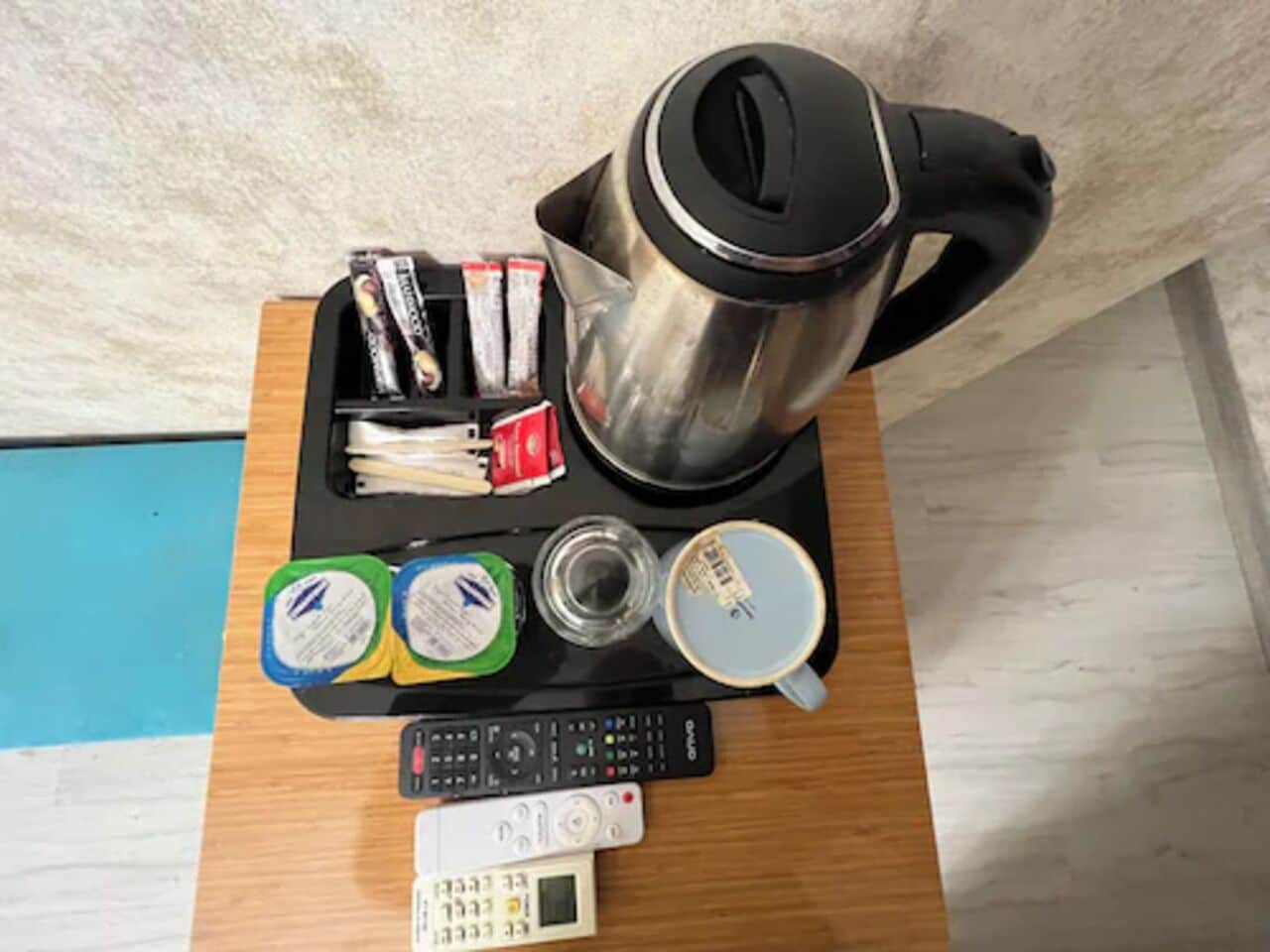 Een houten tafel met een waterkoker, afstandsbedieningen, een kopje en wat etenswaren.
