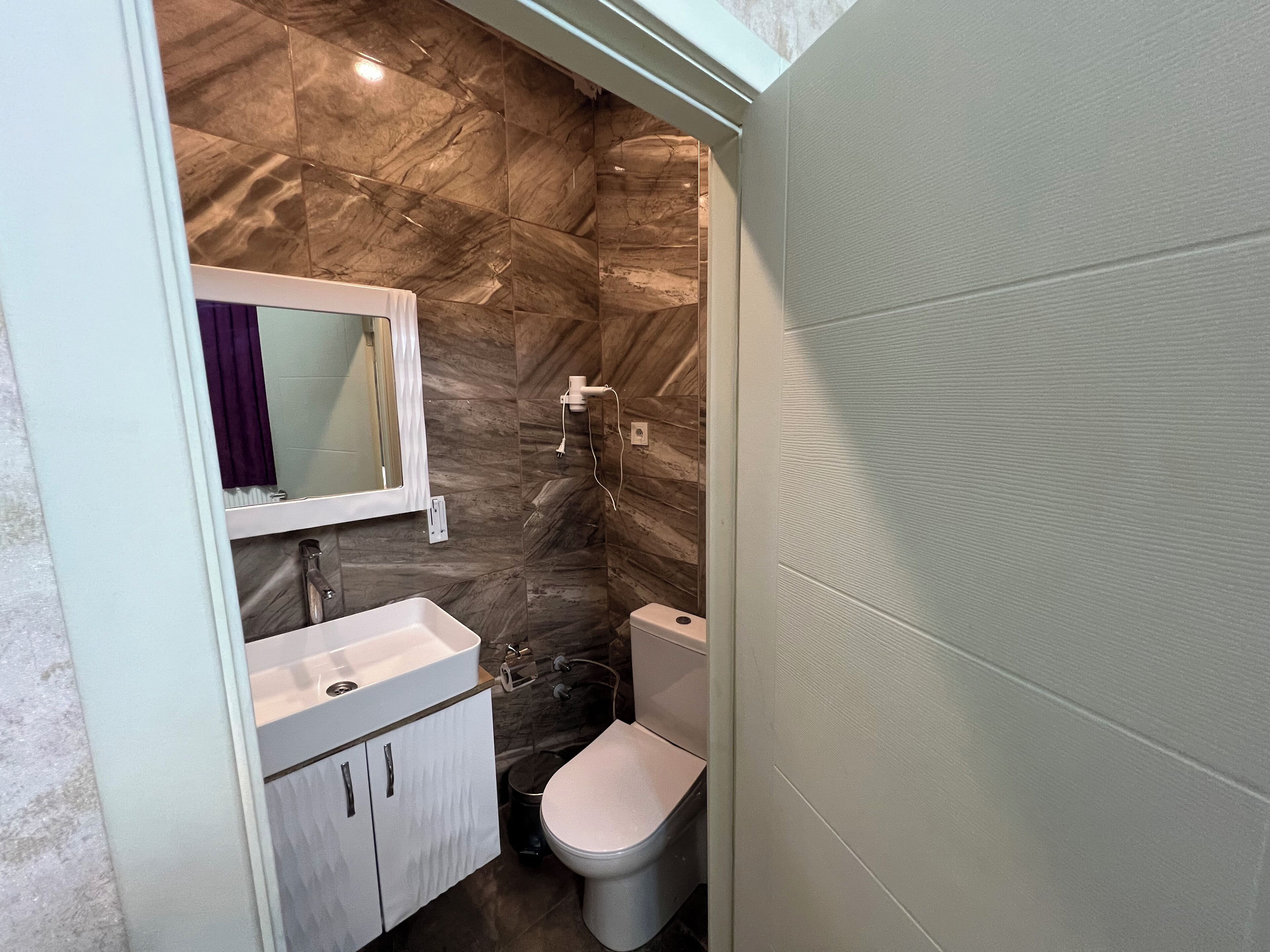 A compact bathroom with a marble backsplash, a white sink, a mirror, and a toilet.