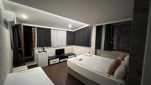 Quarto de hotel com duas camas, uma mesa com computador, um frigorífico pequeno e uma janela com cortinas.