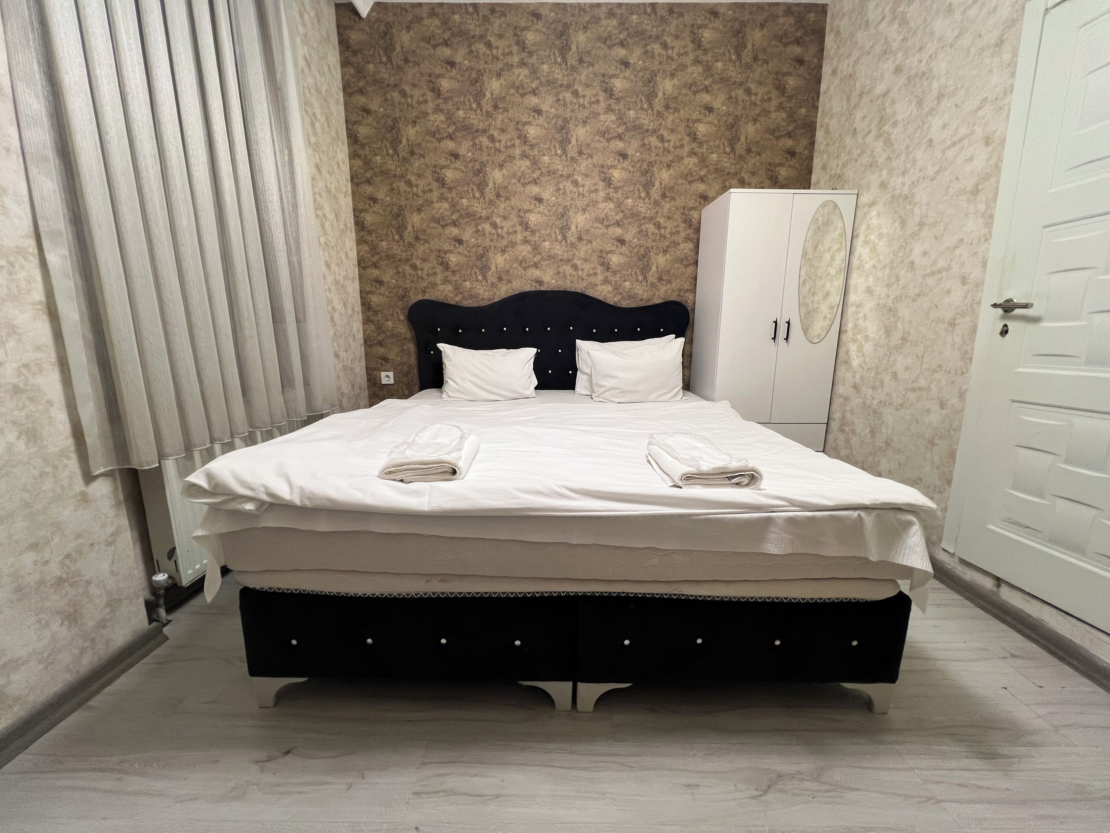 Un lit bien fait, avec du linge de lit et des serviettes blanches, une tête de lit sombre et une armoire adossée à un mur à la texture particulière.