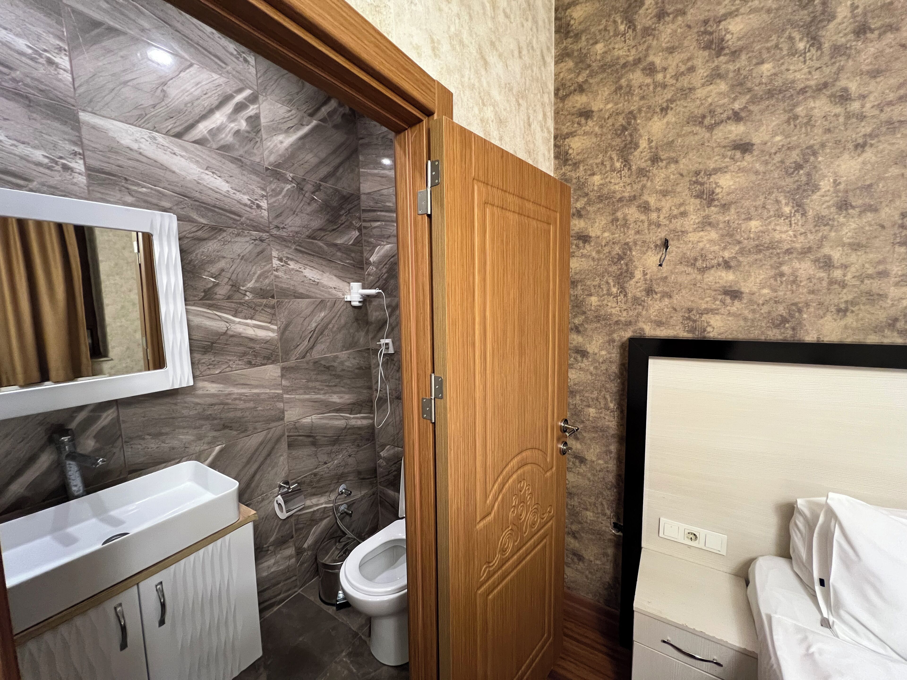 A bathroom with a marble wall, a wooden door, a white sink, a toilet, and a mirror.