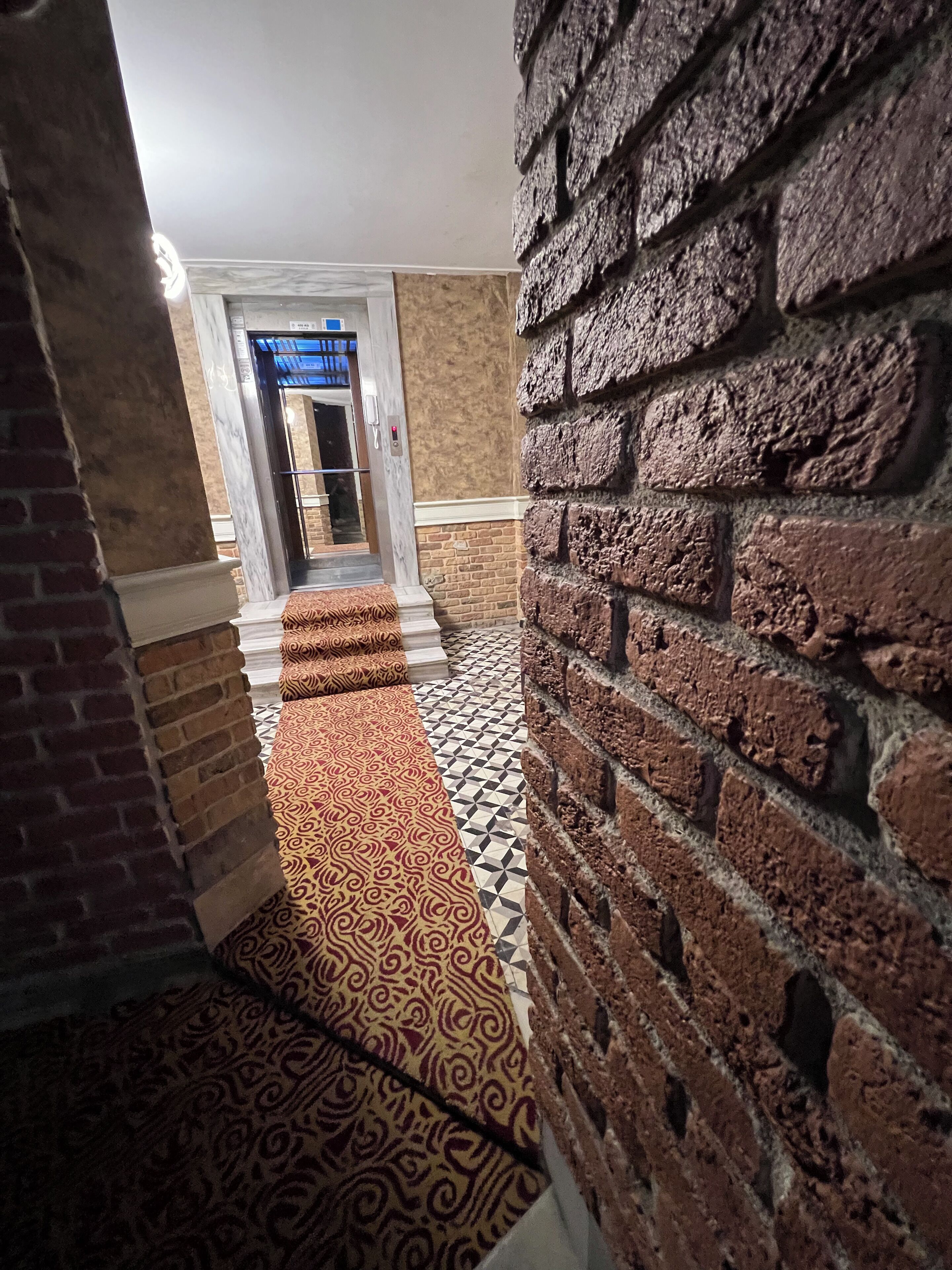 A hallway with patterned carpet, brick walls, and a tiled floor leading to a door with a blue awning.