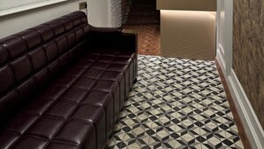 Um corredor com um banco de couro marrom escuro, parede de tijolos brancos, piso estampado e área de cozinha com bancada branca e painel de madeira.