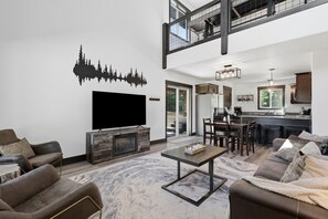 A modern living room with a fireplace, a flat-screen TV, and a dining area with a kitchen in the background.