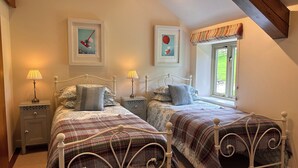 A bedroom with two beds, each with a white metal frame and a plaid blanket. There are bedside tables with lamps, and the room has a sloped ceiling with exposed beams.