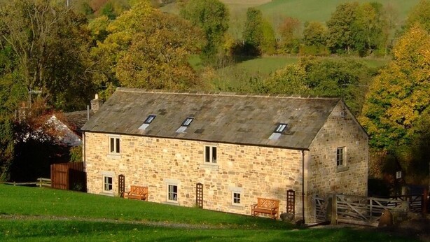 A stone farmhouse with a sloped roof, multiple windows, and a wooden fence.