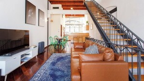 A modern living room with a brown leather sofa, a TV, and a blue patterned rug. A staircase with a metal railing leads to an upper level. - Located opposite Bathers Beach it is just a step away from the beach and cafes. Magnificent Rooftop balcony to enjoy the famous WA Sunsets, head out and enjoy a wine in the many bars surrounding the Loft. (Fremantle)