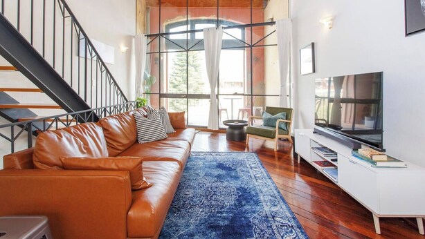 A modern living room with a leather sofa, a blue rug, a TV, and a staircase with metal railings. - Located opposite Bathers Beach it is just a step away from the beach and cafes. Magnificent Rooftop balcony to enjoy the famous WA Sunsets, head out and enjoy a wine in the many bars surrounding the Loft. (Fremantle)