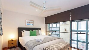 A bedroom with a bed, a nightstand, a ceiling fan, a window with a view of a building, and a framed picture on the wall. - Located opposite Bathers Beach it is just a step away from the beach and cafes. Magnificent Rooftop balcony to enjoy the famous WA Sunsets, head out and enjoy a wine in the many bars surrounding the Loft. (Fremantle)