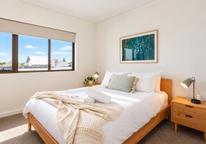 A modern bedroom with a large bed, a nightstand, a bedside lamp, a window with a view, and a framed artwork on the wall.