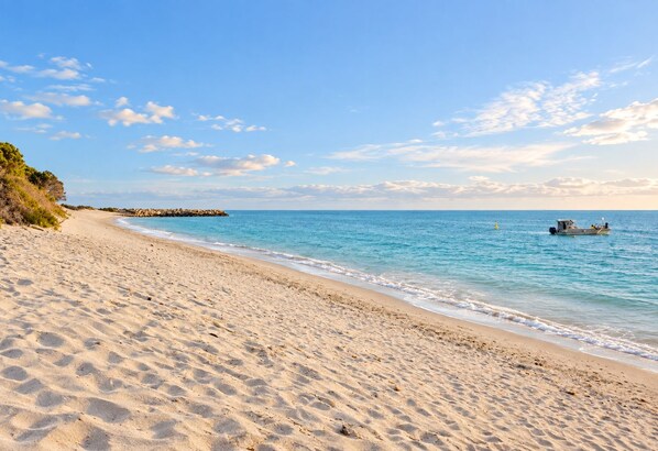 A sandy beach with clear blue water, a rocky outcrop, and a boat in the distance.