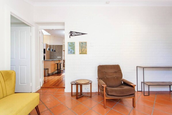 A living room with a brown armchair, a yellow sofa, and a small round table. A kitchen is visible in the background. - This house is a 2 bedroom(s), 2 bathrooms, located in Fremantle, WA. (Fremantle)