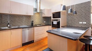 A modern kitchen with wooden cabinets, a stainless steel dishwasher, a black countertop, and a tiled backsplash. - This house is a 2 bedroom(s), 2 bathrooms, located in Fremantle, WA. (Fremantle)