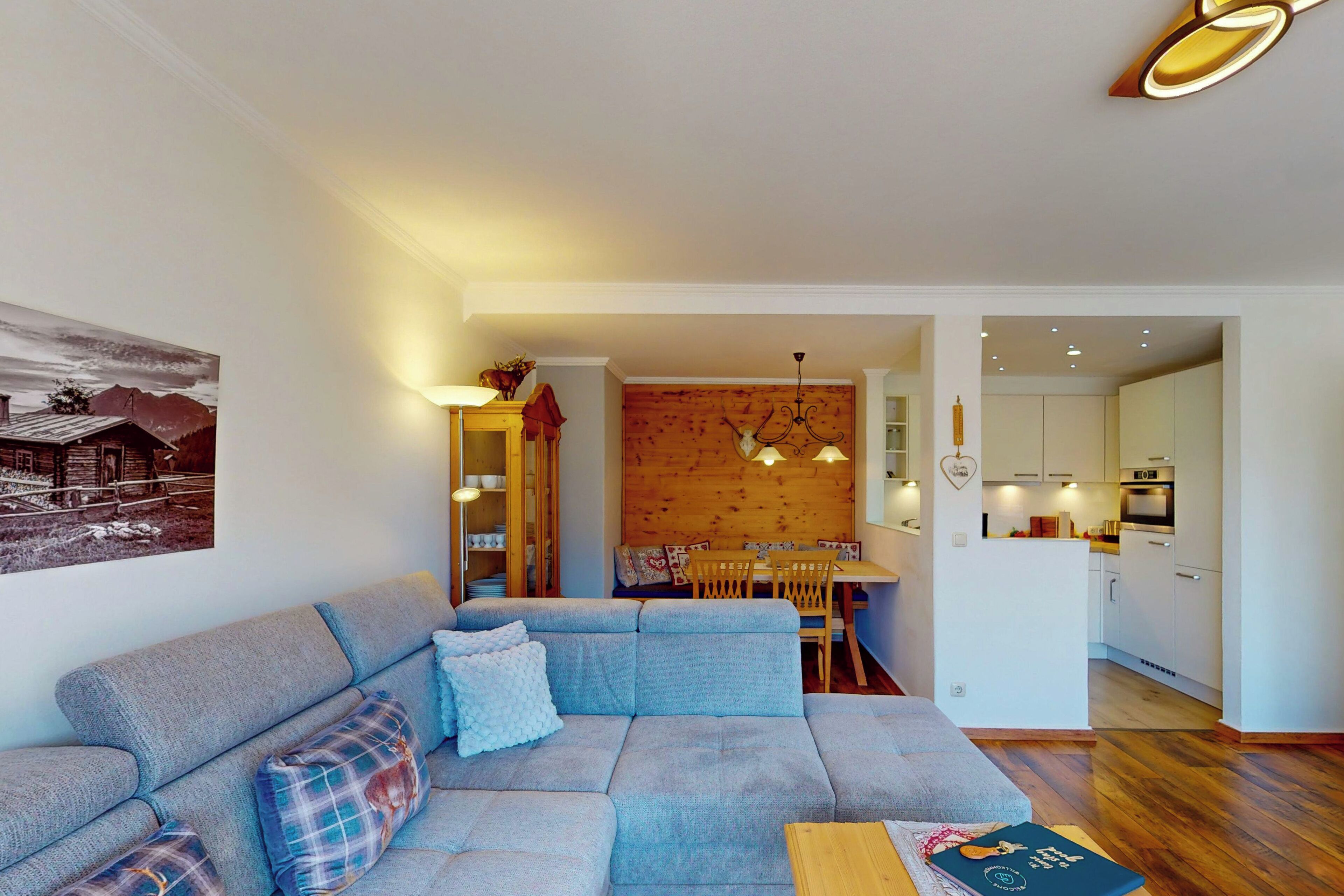 A modern living room with a grey sofa, wooden dining table, and a kitchen in the background.