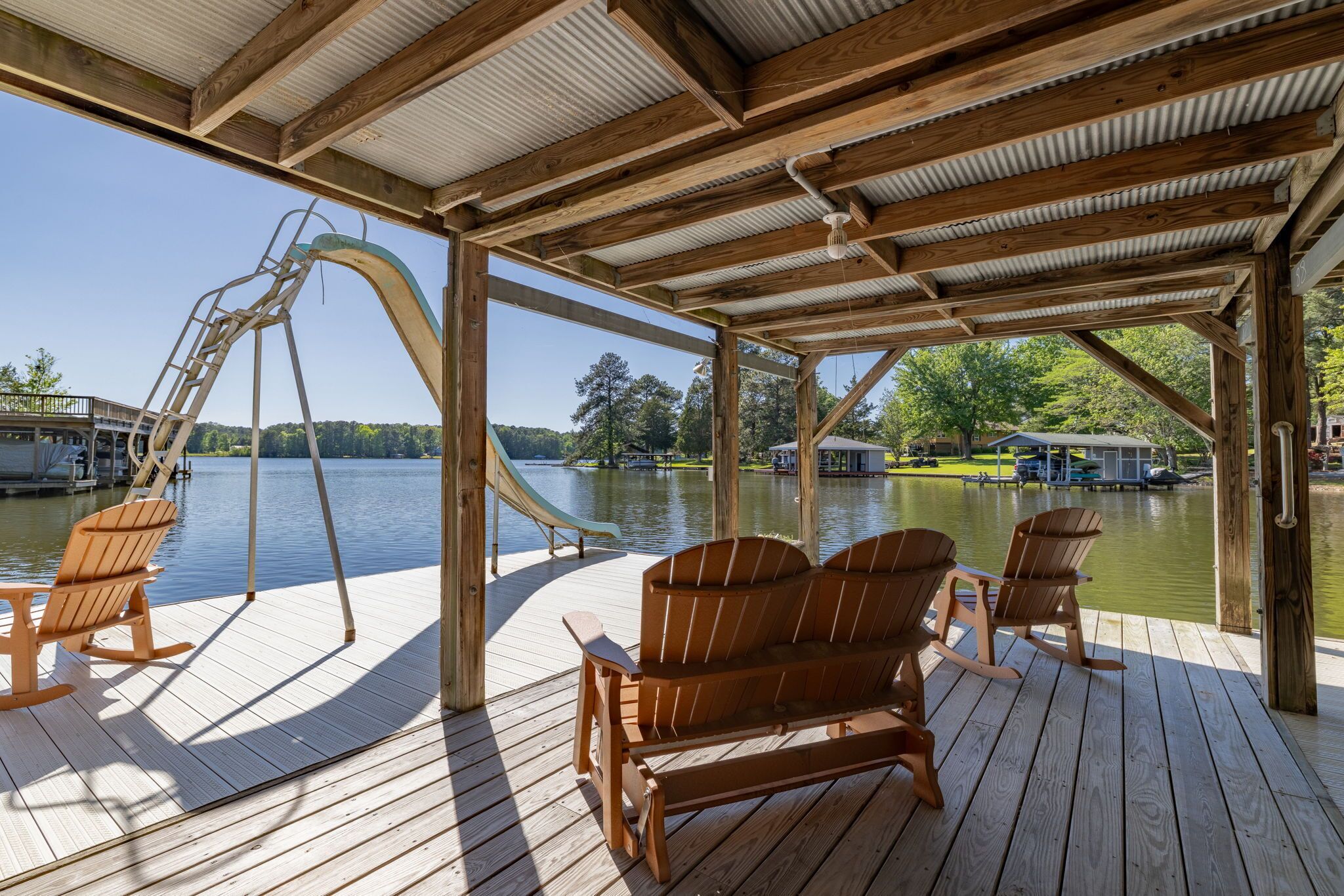 A wooden deck with Adirondack chairs, a slide, and a view of a lake and buildings in the background.
