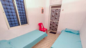 Una habitación pequeña con dos camas individuales, una silla roja, un ventanal con vidrio azulado y una puerta con estampado floral.