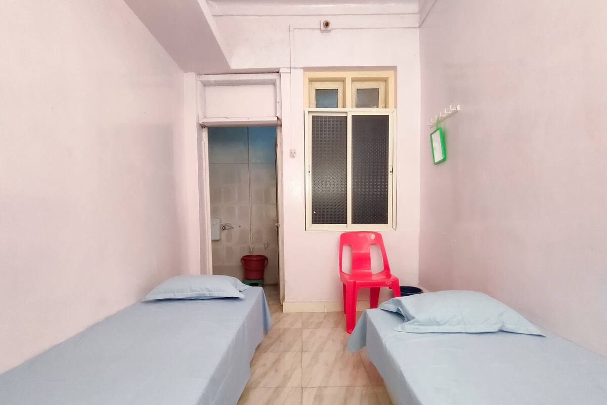A small, narrow room with two single beds, a window with a screen, a red plastic chair, and a bucket on the floor.