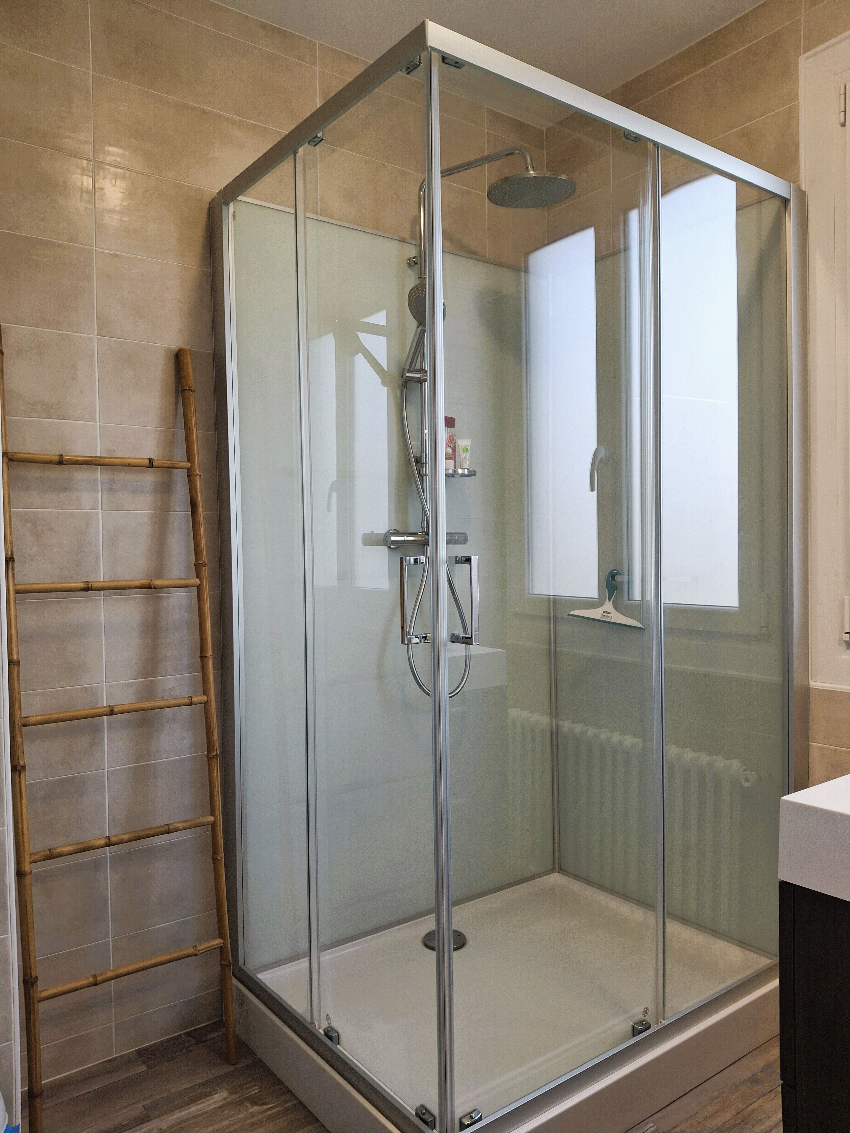 A glass-enclosed shower with a wooden ladder, a towel rack, and a small shelf with toiletries.