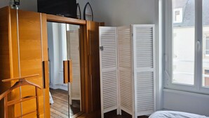 A bedroom with a wooden wardrobe, a television on top, a white bed, and a window with a view of buildings outside. - Warm and Authentic! The watchmaker’s house, downtown Crozon (Crozon)