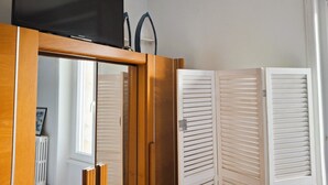 A room with a wooden wardrobe, a television mounted above it, and a window with blinds. - Warm and Authentic! The watchmaker’s house, downtown Crozon (Crozon)