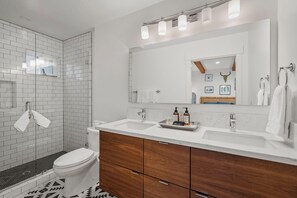 Une salle de bain moderne avec une douche carrelée de blanc, des toilettes blanches, un meuble-lavabo en bois avec un grand miroir et un lavabo blanc.