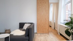 A modern living room with a grey sofa, a white furry throw, a wooden partition, and a view of a brick building through a window. - Beautiful apartment in Liège center - 1st floor (Liège)