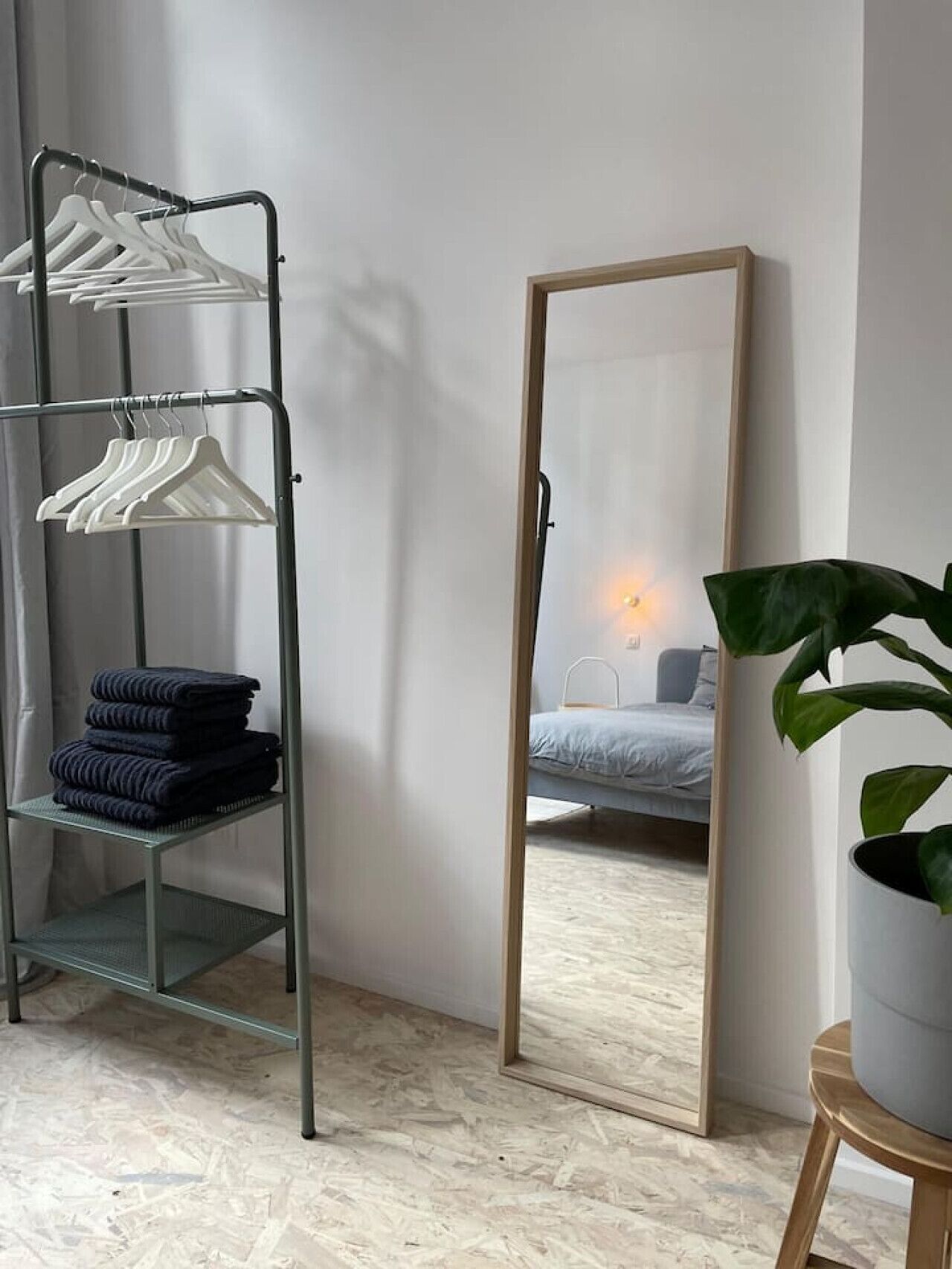 Une pièce avec une étagère en métal où sont rangés des cintres blancs et des vêtements pliés, un grand miroir qui reflète un lit et une lampe, et une plante en pot.