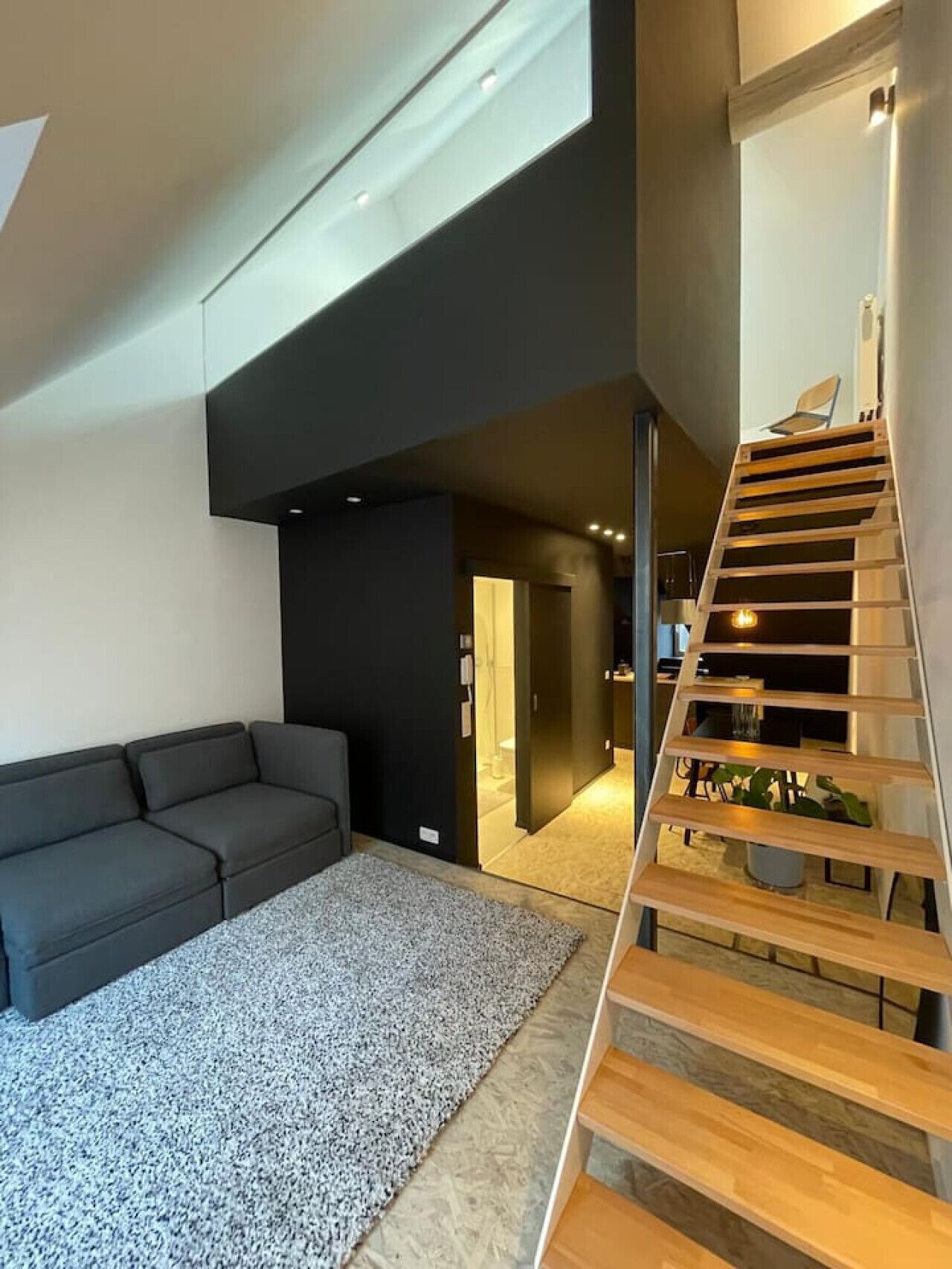 A modern living room with a dark sofa, a patterned rug, and a wooden staircase leading to an upper level.