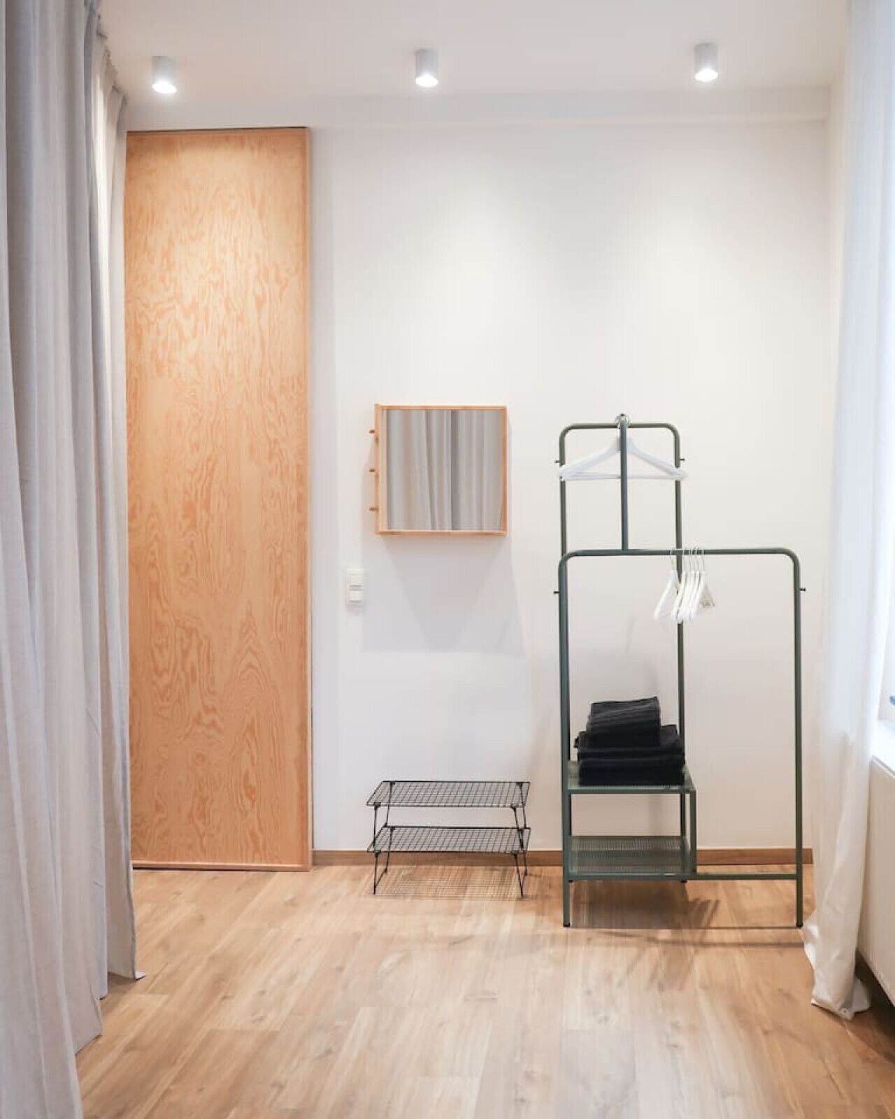 Una habitación con una puerta de madera, un perchero metálico con ropa, un estante pequeño y un espejo en la pared.