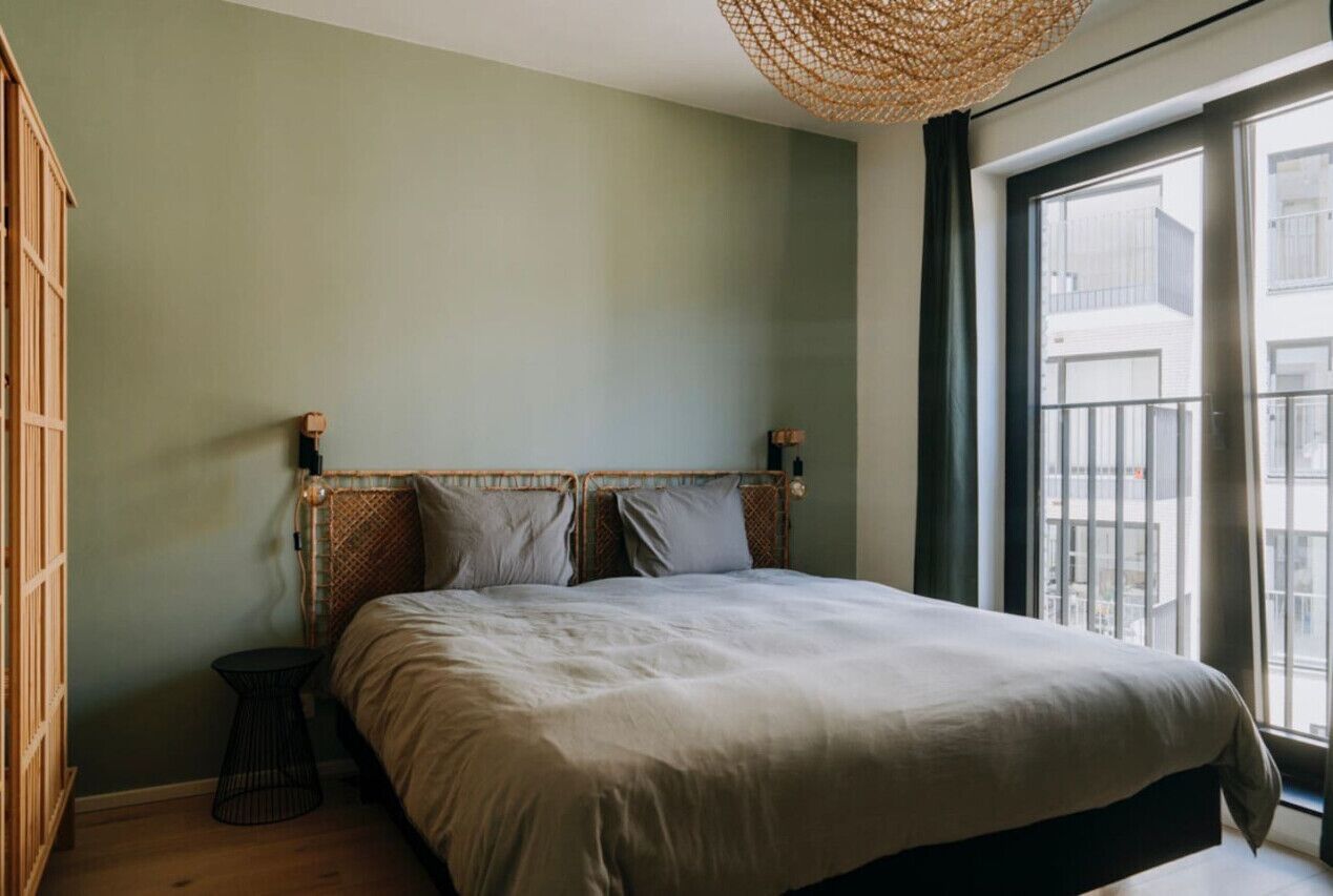 Une chambre avec un grand lit, une petite table de chevet, une tête de lit en osier et une vue sur l’extérieur par une fenêtre.