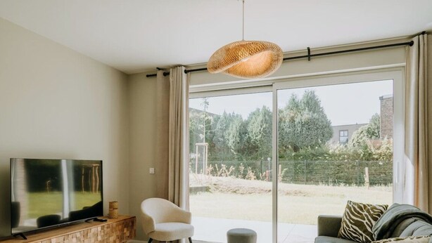 Ein modernes Wohnzimmer mit einem Flachbildfernseher, einem weißen Sessel, einer Couch mit einem Kissen mit ZEBRA-MUSTER und einem großen Fenster mit Blick auf einen Garten und Bäume.