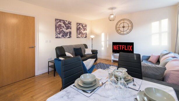 Un salon moderne avec un coin repas préparé pour un repas, un canapé, un téléviseur affichant « NETFLIX » et des œuvres d’art accrochées aux murs.