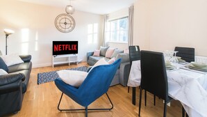 Un salon moderne avec un canapé, un fauteuil bleu, une table à manger dressée pour deux personnes, un téléviseur affichant le logo « NETFLIX » et une horloge au mur.