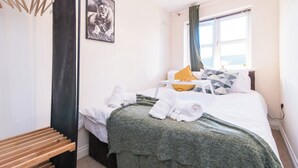Une chambre d’hôtel avec un lit, une couverture verte, des serviettes blanches, un banc en bois, un tableau et une fenêtre avec des rideaux.