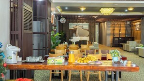 A dining area with a wooden table, chairs, a coffee station, and a buffet with fruits and a juice pitcher. - Lucky City Hotel (Vientiane)