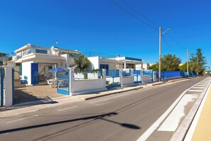 Uma fileira de edifícios modernos, com dois andares, portões azuis e paredes brancas, céu limpo e rua organizada.